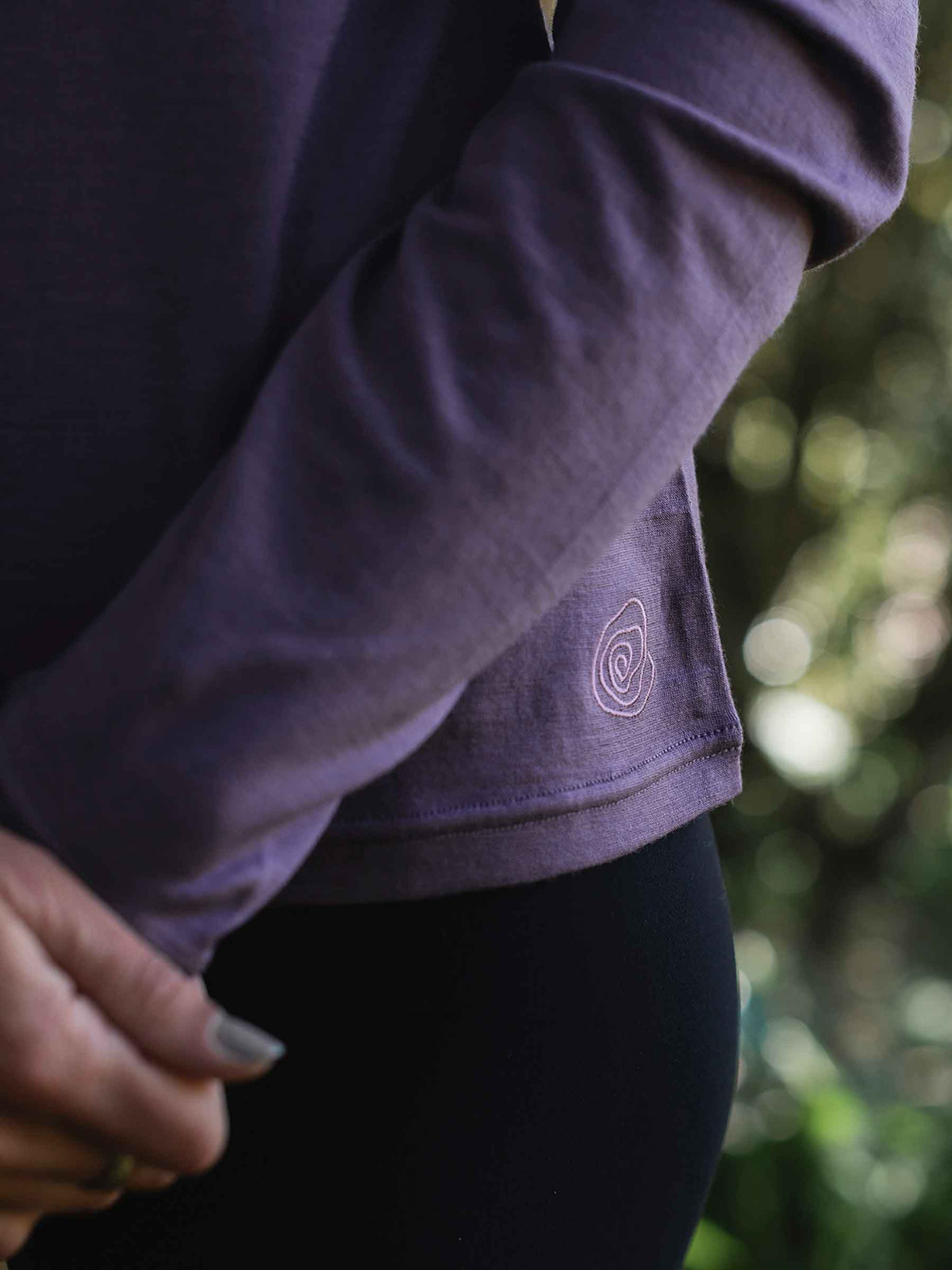 Person wearing a purple long-sleeve shirt with a logo, standing outdoors with blurred greenery in the background.