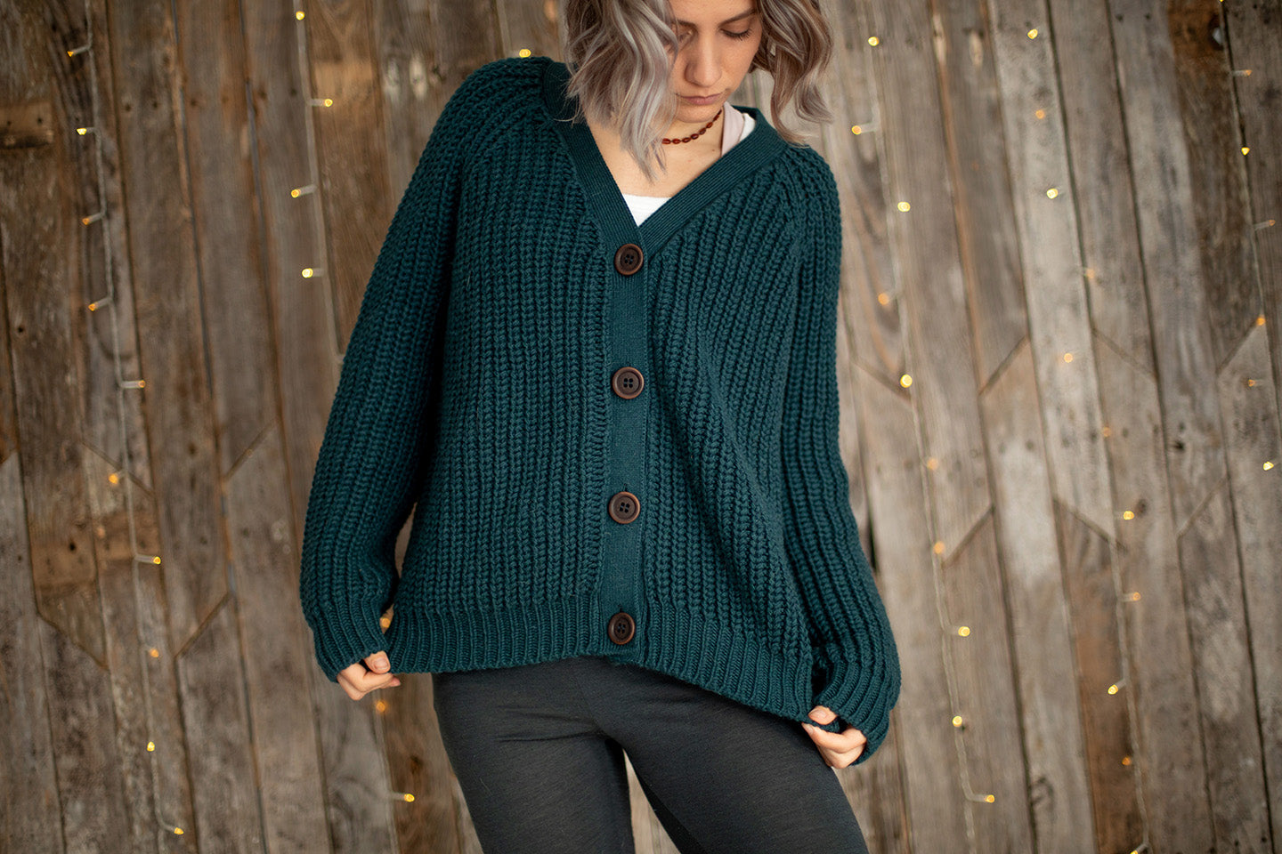 Nui Organics Women's Bernard Cardigan in Deep Sea — woman in chunky teal merino cardigan with corozo buttons and grey leggings against a wooden wall with fairy lights