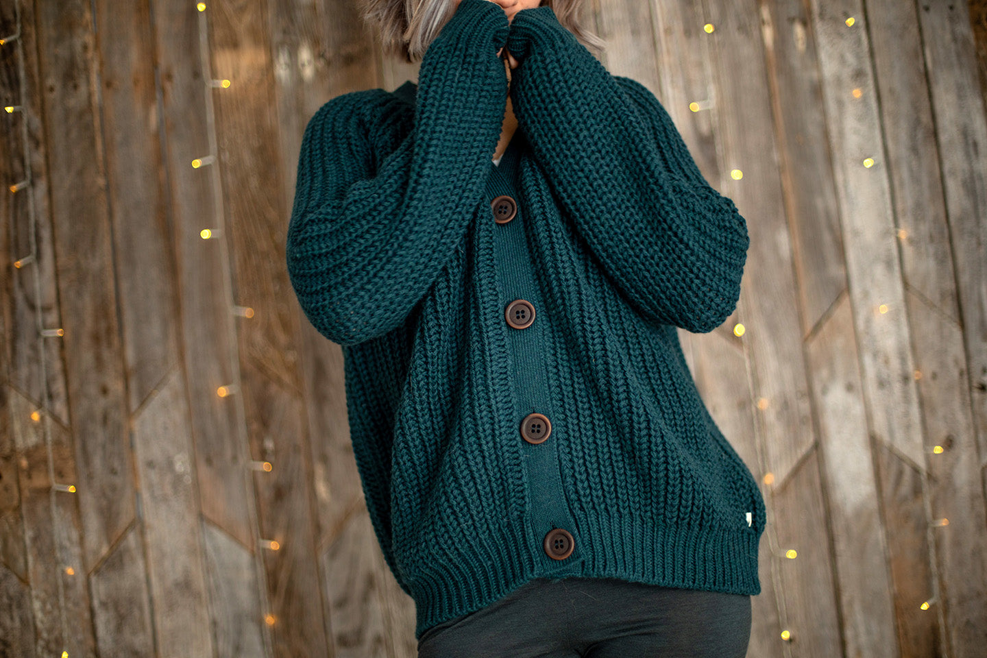 Nui Organics Women's Bernard Cardigan in Deep Sea — close-up of chunky teal merino knit and corozo nut buttons against a wooden backdrop with fairy lights