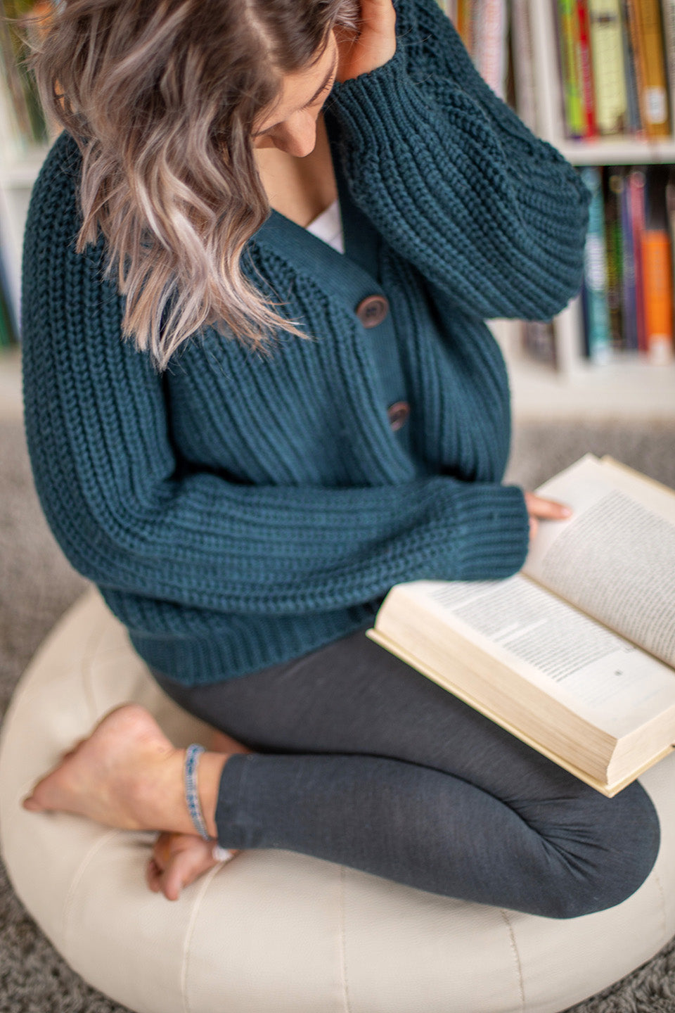 Nui Organics Women's Bernard Cardigan in Deep Sea — overhead view of woman in teal chunky merino cardigan seated cross-legged reading a book
