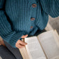 Nui Organics Women's Bernard Cardigan in Deep Sea — close-up of corozo nut button detail on chunky teal merino cardigan with open book in lap