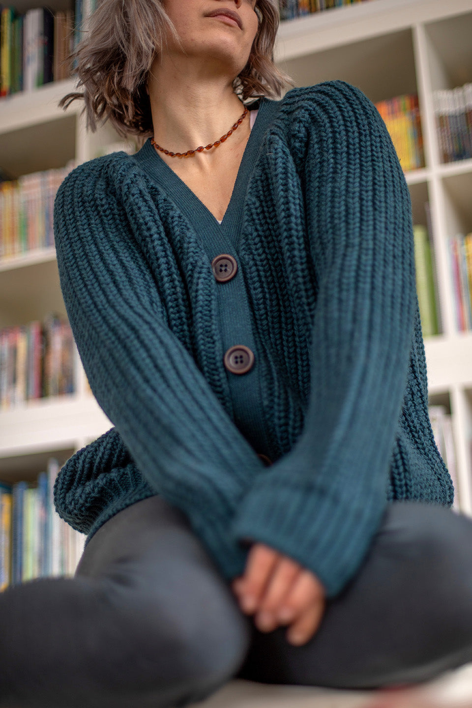 Nui Organics Women's Bernard Cardigan in Deep Sea — close-up of v-neck and corozo button detail on chunky teal merino wool cardigan, seated indoors