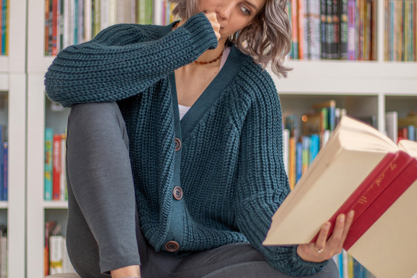 Nui Organics Women's Bernard Cardigan in Deep Sea — woman in chunky teal merino cardigan reading a book in front of a bookshelf