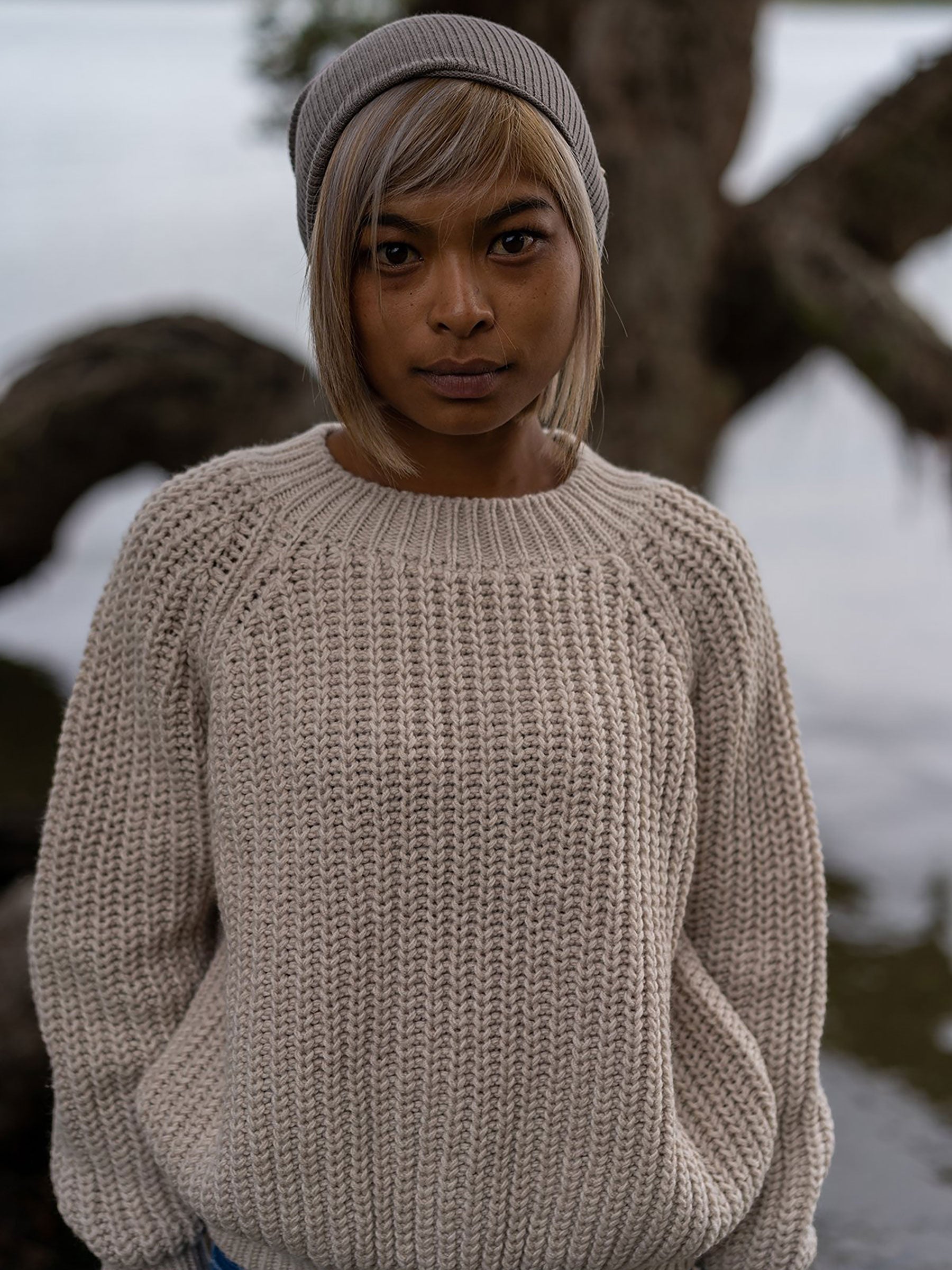Nui Organics Women's Fishline Sweater in Oatmeal — woman wearing oatmeal chunky merino sweater and grey beanie beside a coastal tree