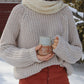 Nui Organics Women's Fishline Sweater in Oatmeal — close-up of chunky merino knit sweater worn with cream snood, holding a coffee mug in the snow