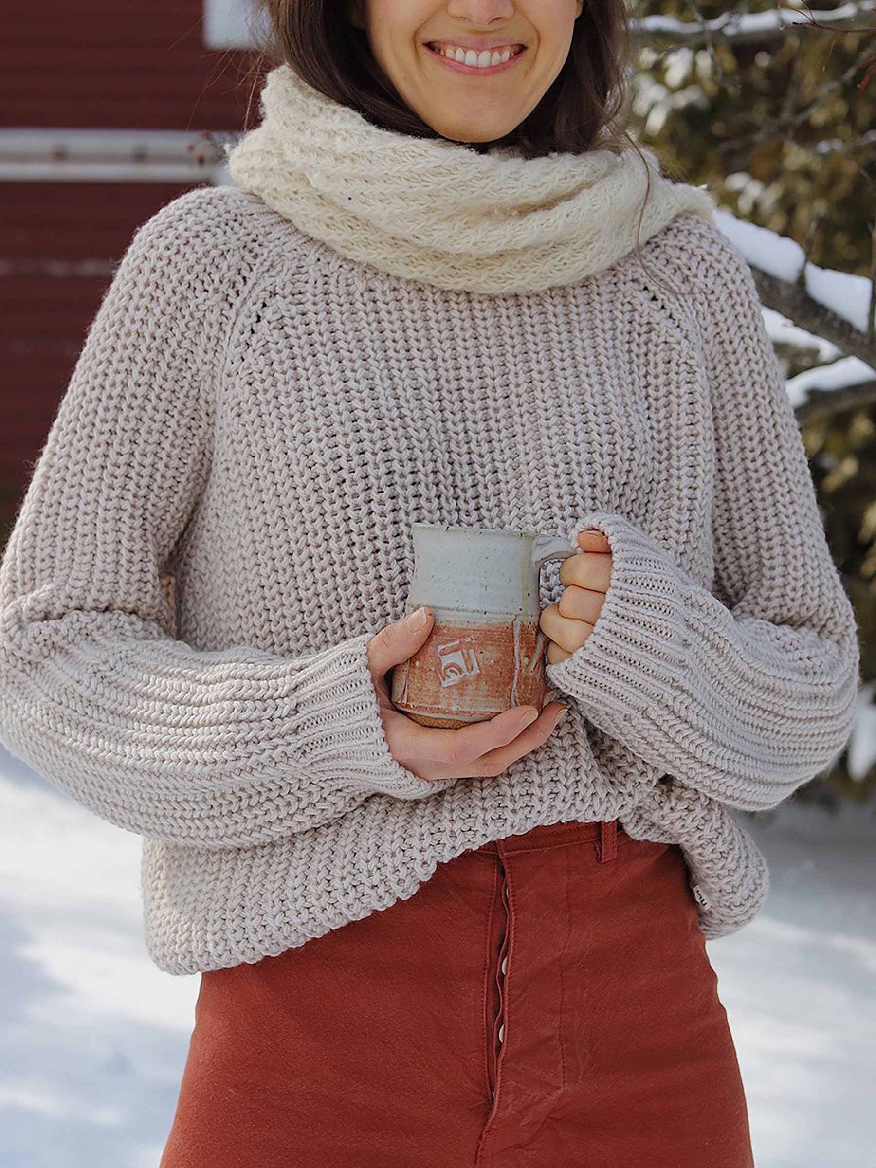 Nui Organics Women's Fishline Sweater in Oatmeal — close-up of chunky merino knit sweater worn with cream snood, holding a coffee mug in the snow