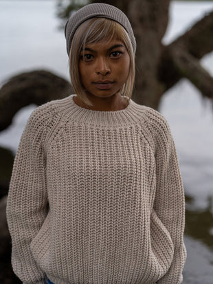 Nui Organics Women's Fishline Sweater in Oatmeal — woman wearing oatmeal chunky merino sweater and grey beanie beside a coastal tree