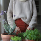 Nui Organics Women's Fishline Sweater in Oatmeal — woman in chunky merino knit sweater and rust trousers tending herbs indoors