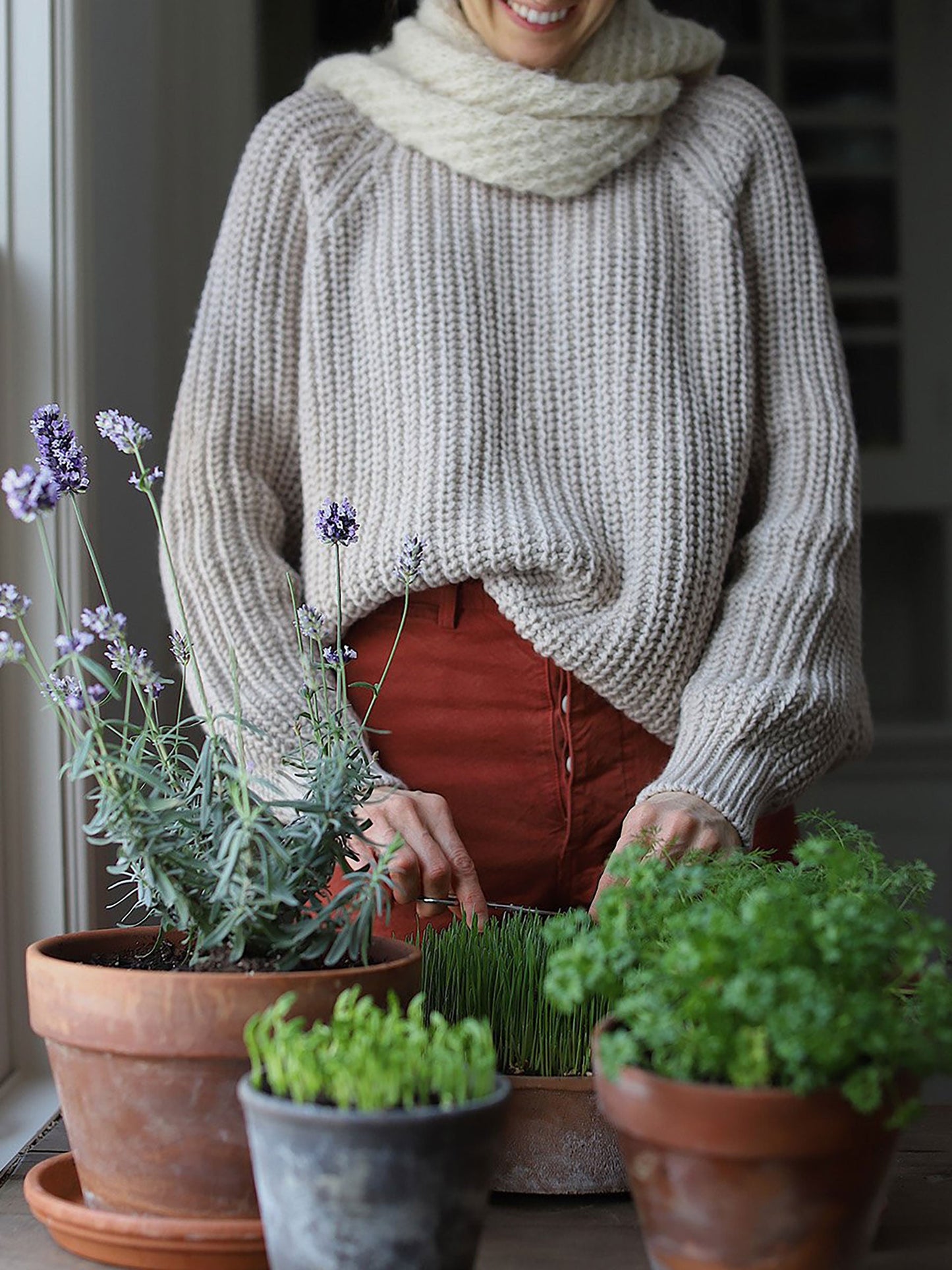 Nui Organics Women's Fishline Sweater in Oatmeal — woman in chunky merino knit sweater and rust trousers tending herbs indoors