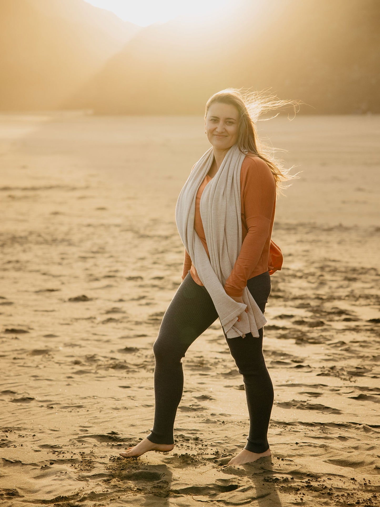 Nui Organics Women's Knit Leggings in Charcoal — woman wearing charcoal merino leggings with rust top and natural wrap on a sandy beach at sunset