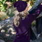 Nui Organics Women's Hopu Top in Plum — rear view of woman in plum rib knit merino top and matching beanie beside a coastal tree