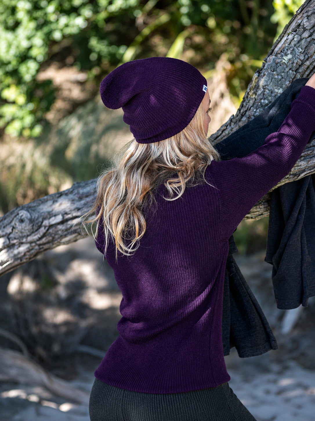 Nui Organics Women's Hopu Top in Plum — rear view of woman in plum rib knit merino top and matching beanie beside a coastal tree