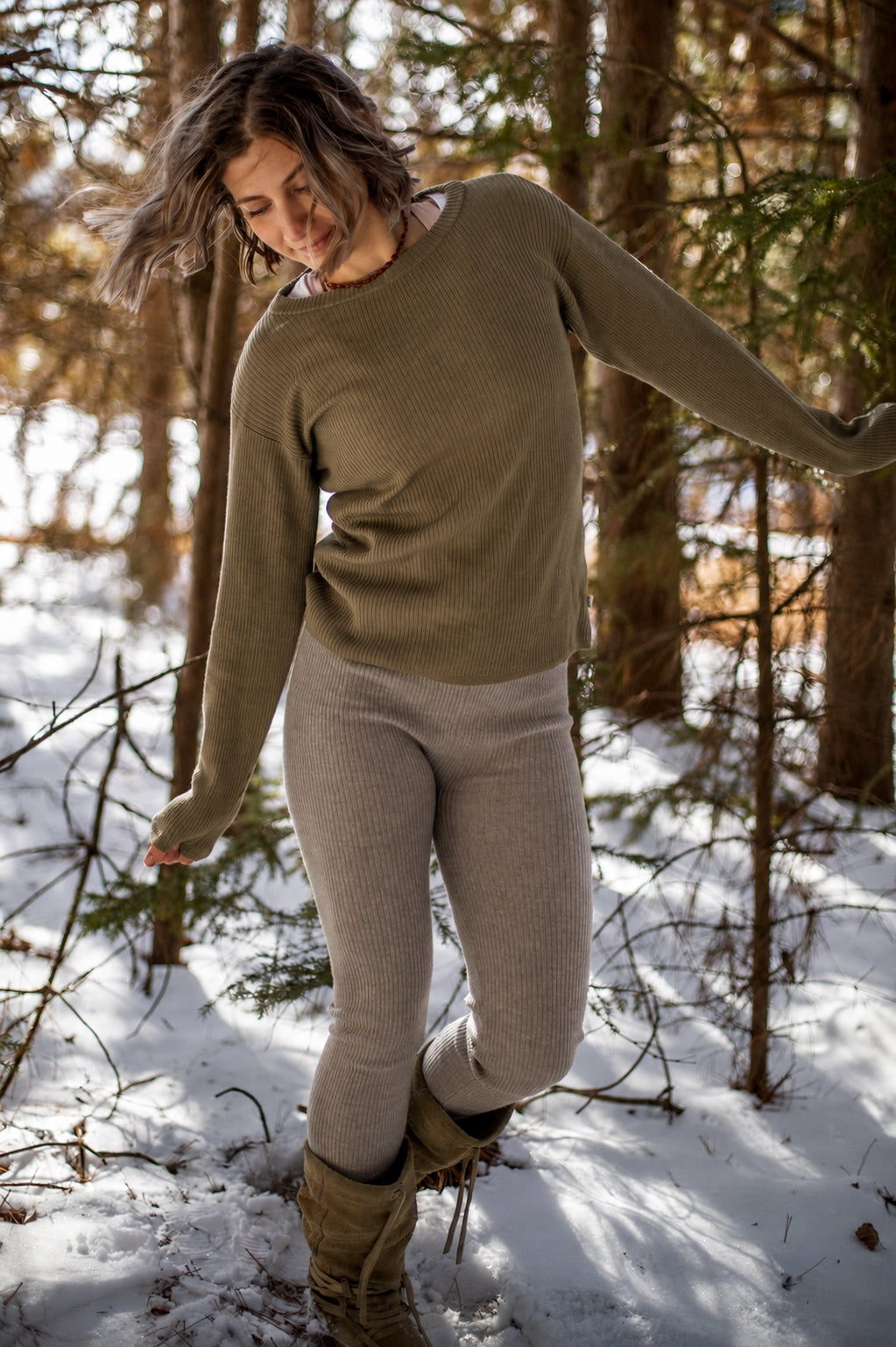Nui Organics Women's Knit Leggings in Sparrow — woman in sparrow rib knit merino leggings and olive sweater walking through a snowy forest.