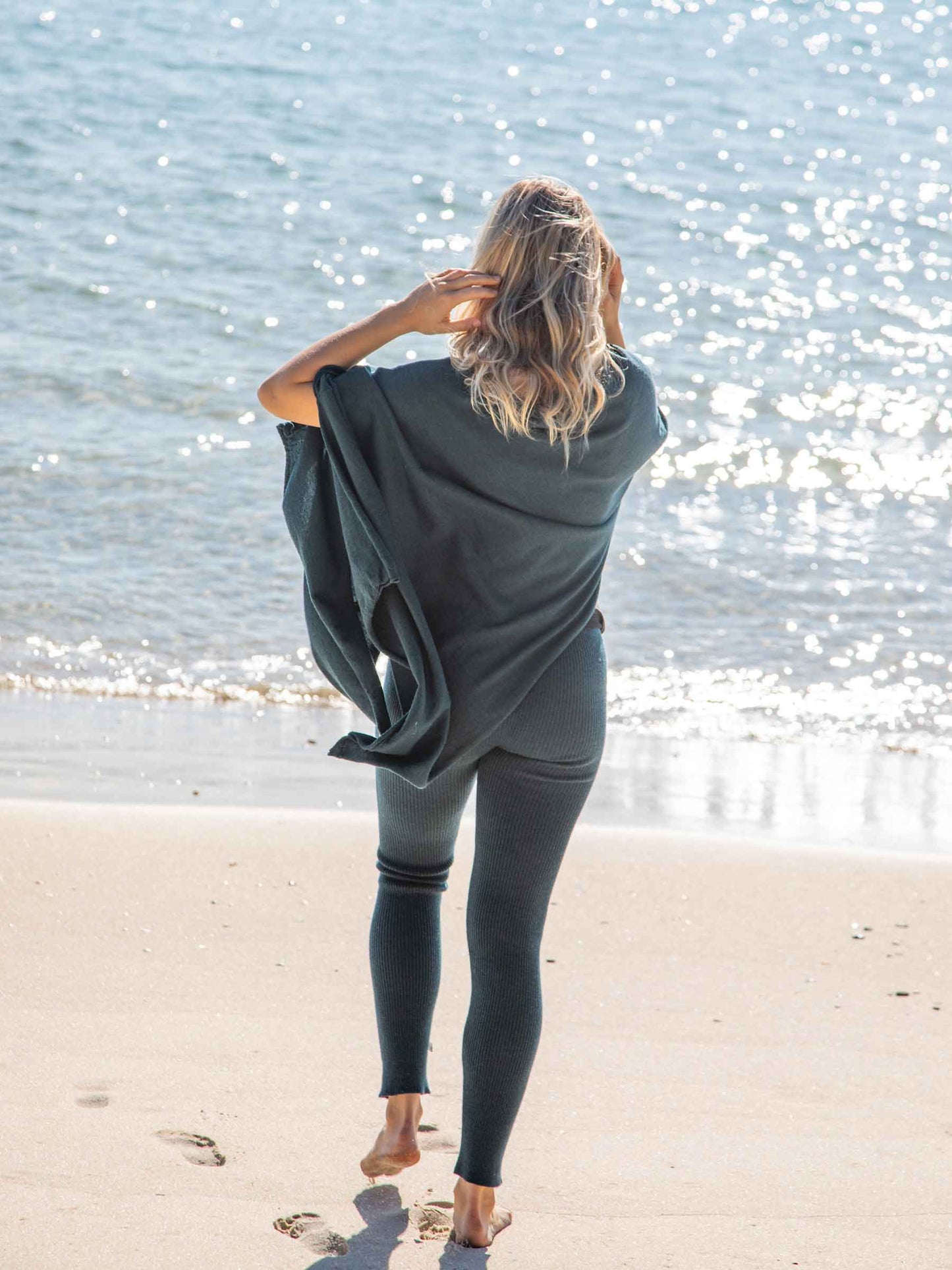 Nui Organics Women's Knit Leggings in Teal — rear view of woman in teal rib knit merino leggings and poncho walking barefoot toward the ocean