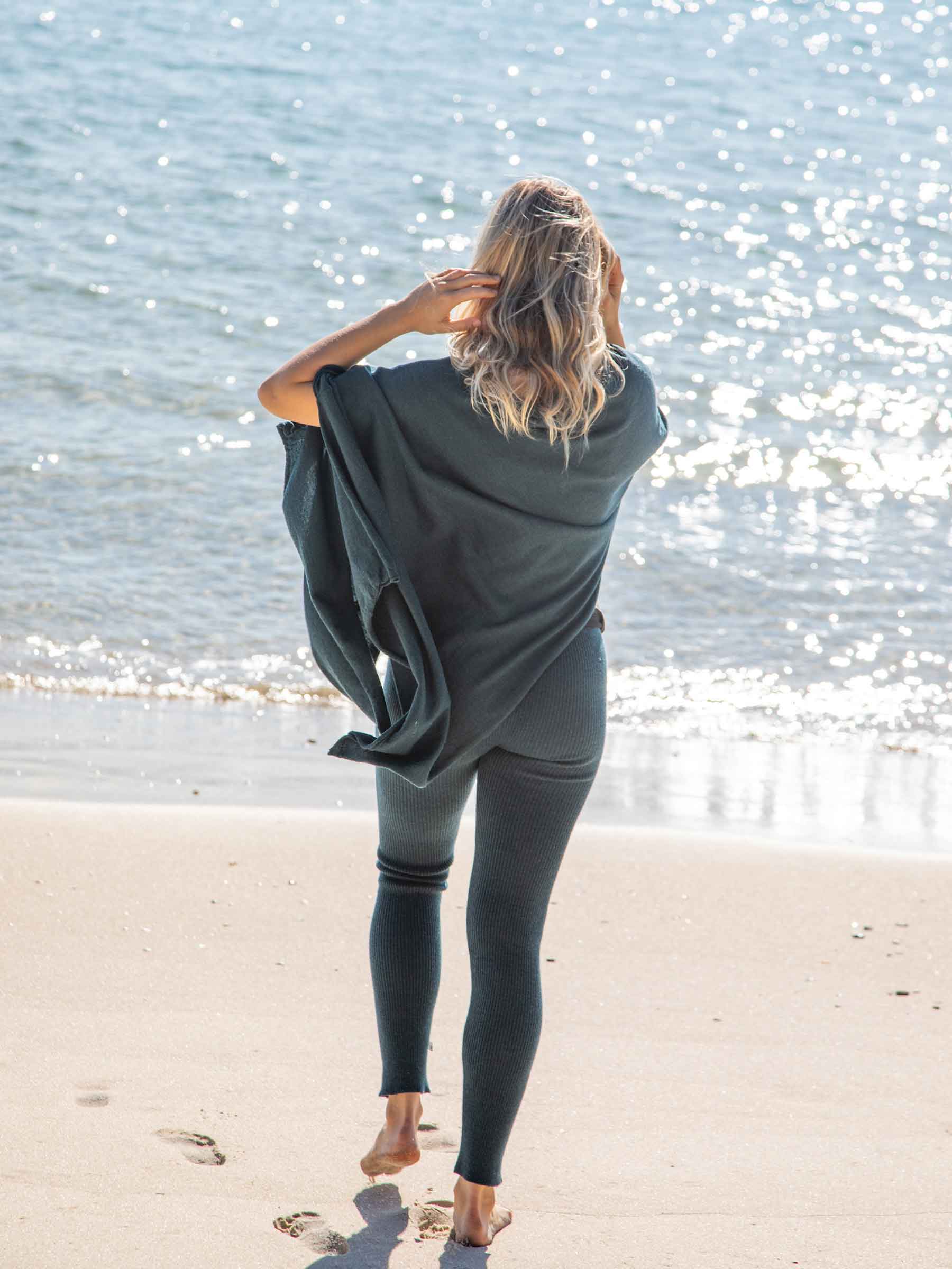 Nui Organics Women's Knit Leggings in Teal — rear view of woman in teal rib knit merino leggings and poncho walking barefoot toward the ocean