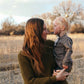 Nui Organics Women's Fishline Sweater in Kelp — woman in chunky olive merino knit sweater holding a young child in a golden winter field