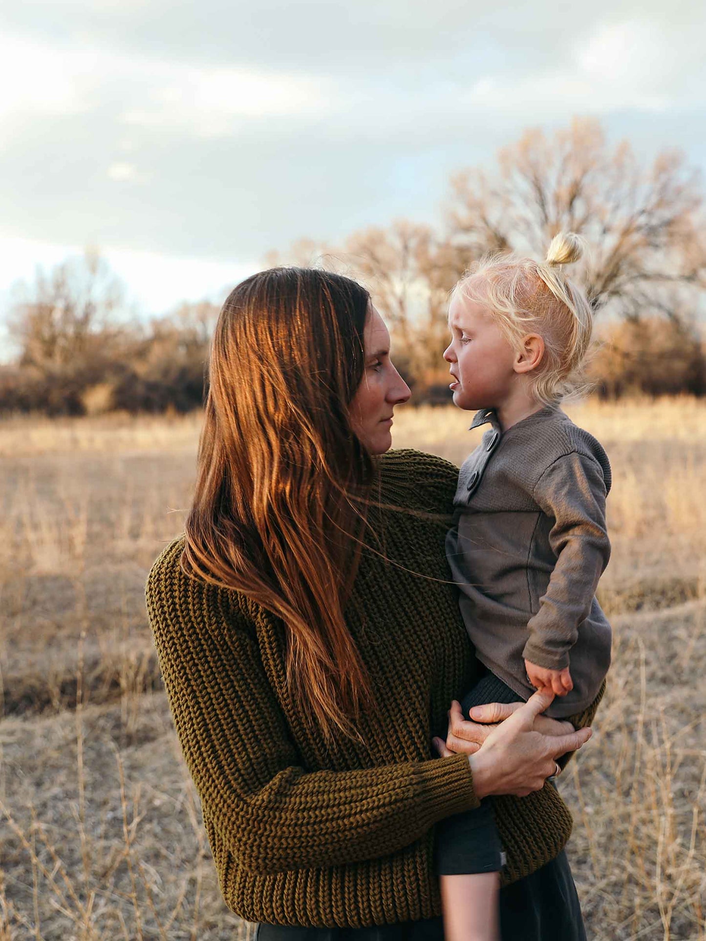 Nui Organics Women's Fishline Sweater in Kelp — woman in chunky olive merino knit sweater holding a young child in a golden winter field