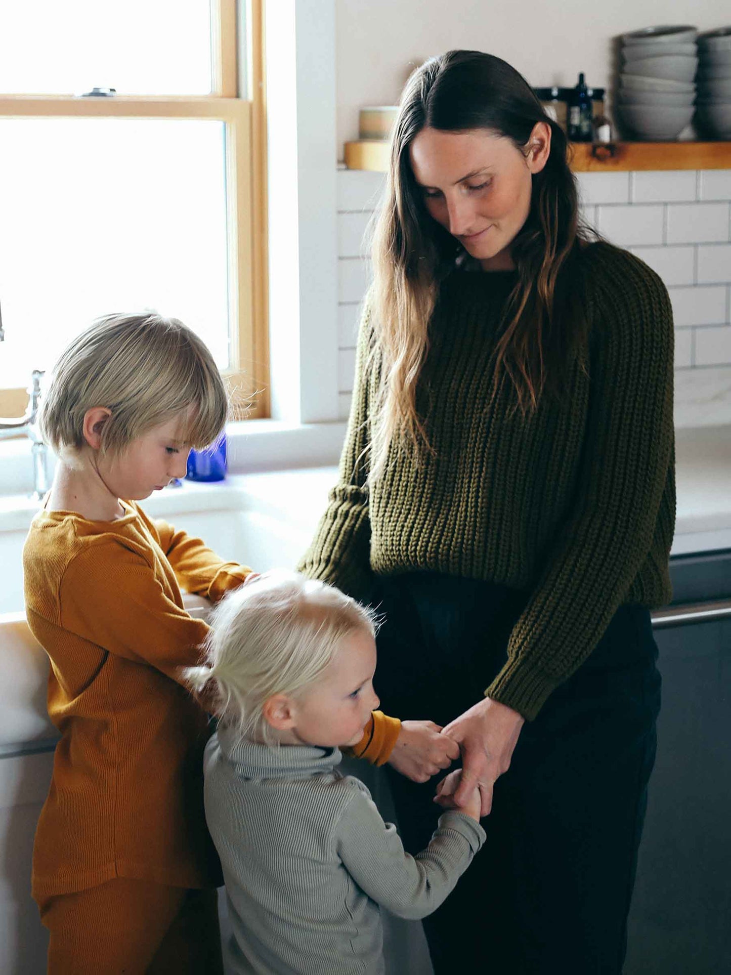 Nui Organics Women's Fishline Sweater in Kelp — woman in chunky olive merino knit sweater with two young children in a home kitchen