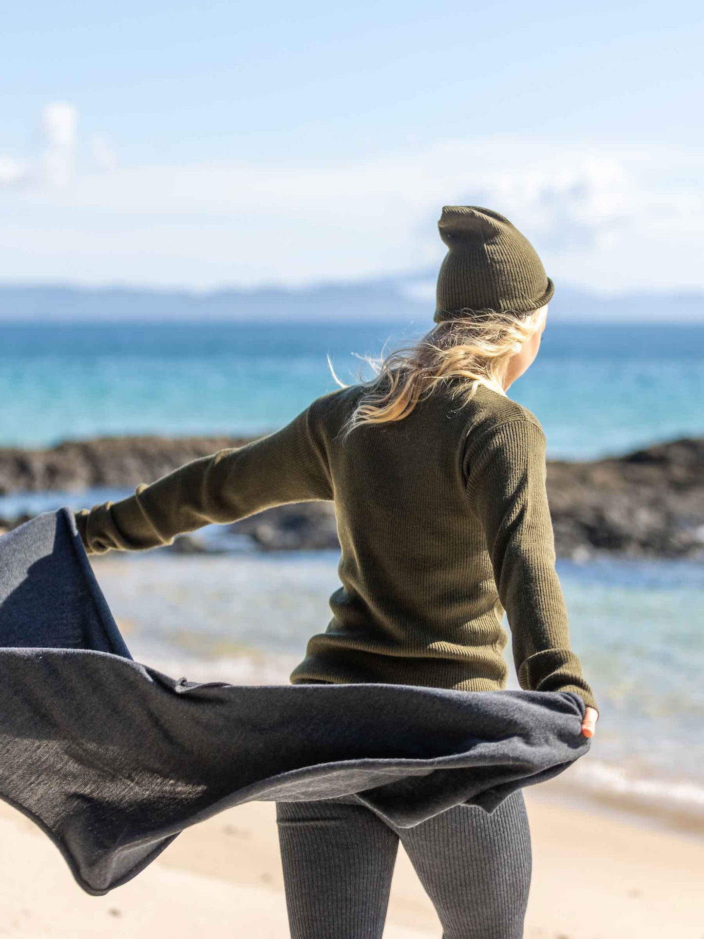Nui Organics Women's Knit Leggings in Charcoal — rear view of woman in charcoal rib knit merino leggings and olive top holding a wrap on a breezy beach
