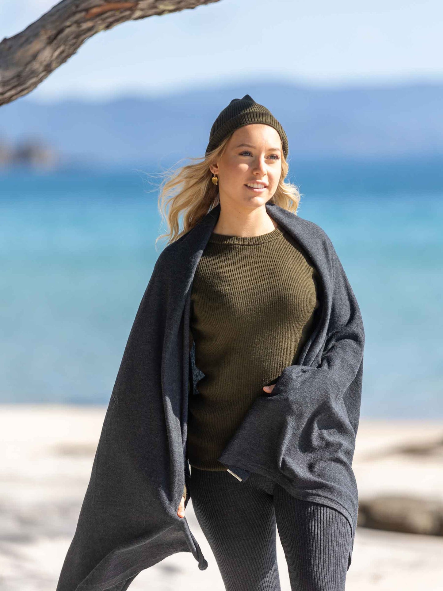 Nui Organics Women's Knit Leggings in Charcoal — woman in charcoal merino leggings, olive knit top and beanie wrapped in a blanket on a sunny beach
