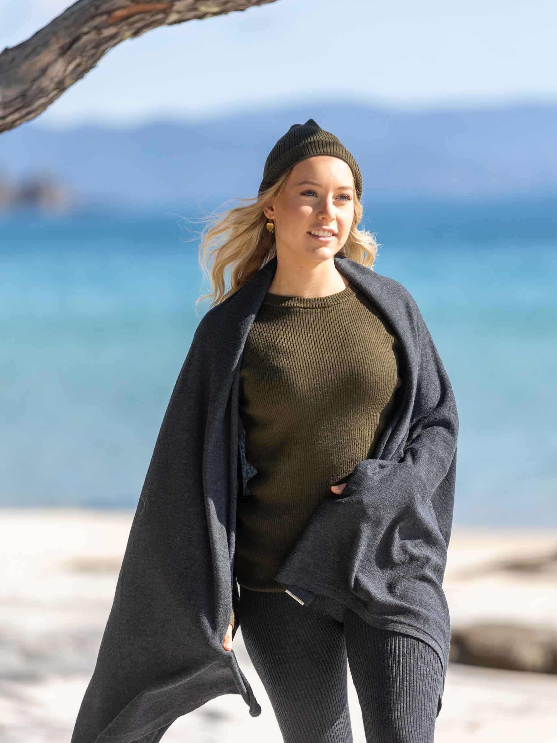 Nui Organics Women's Knit Leggings in Charcoal — woman in charcoal merino leggings, olive knit top and beanie wrapped in a blanket on a sunny beach