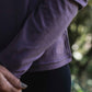 Person wearing a purple long-sleeve shirt with a logo, standing outdoors with blurred greenery in the background.