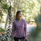 Woman in a purple long-sleeve shirt standing in a forested area with trees and plants around her.
