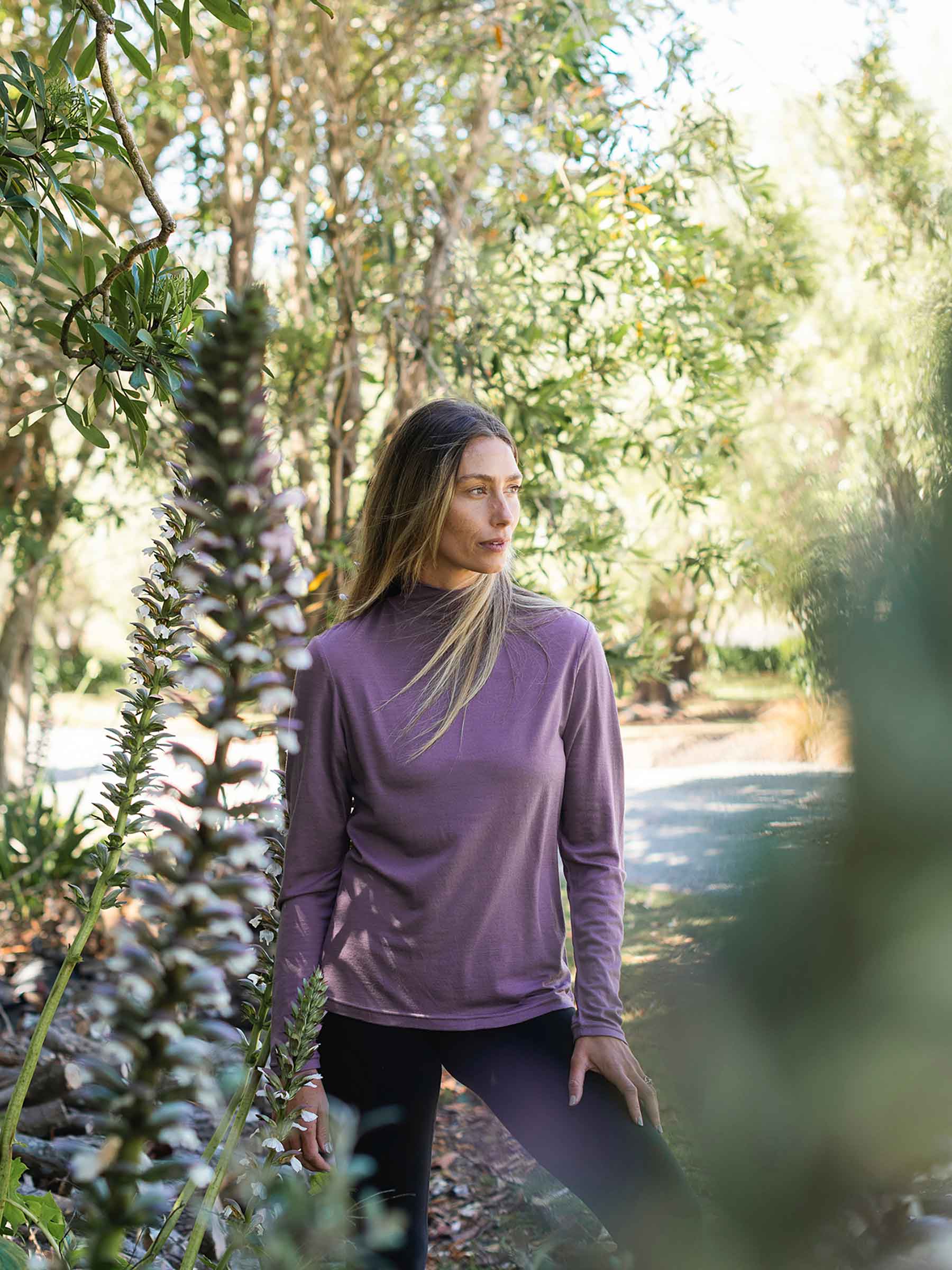Woman in a purple long-sleeve shirt standing in a forested area with trees and plants around her.