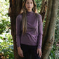Woman wearing a purple long-sleeve top standing between two trees in a forest.