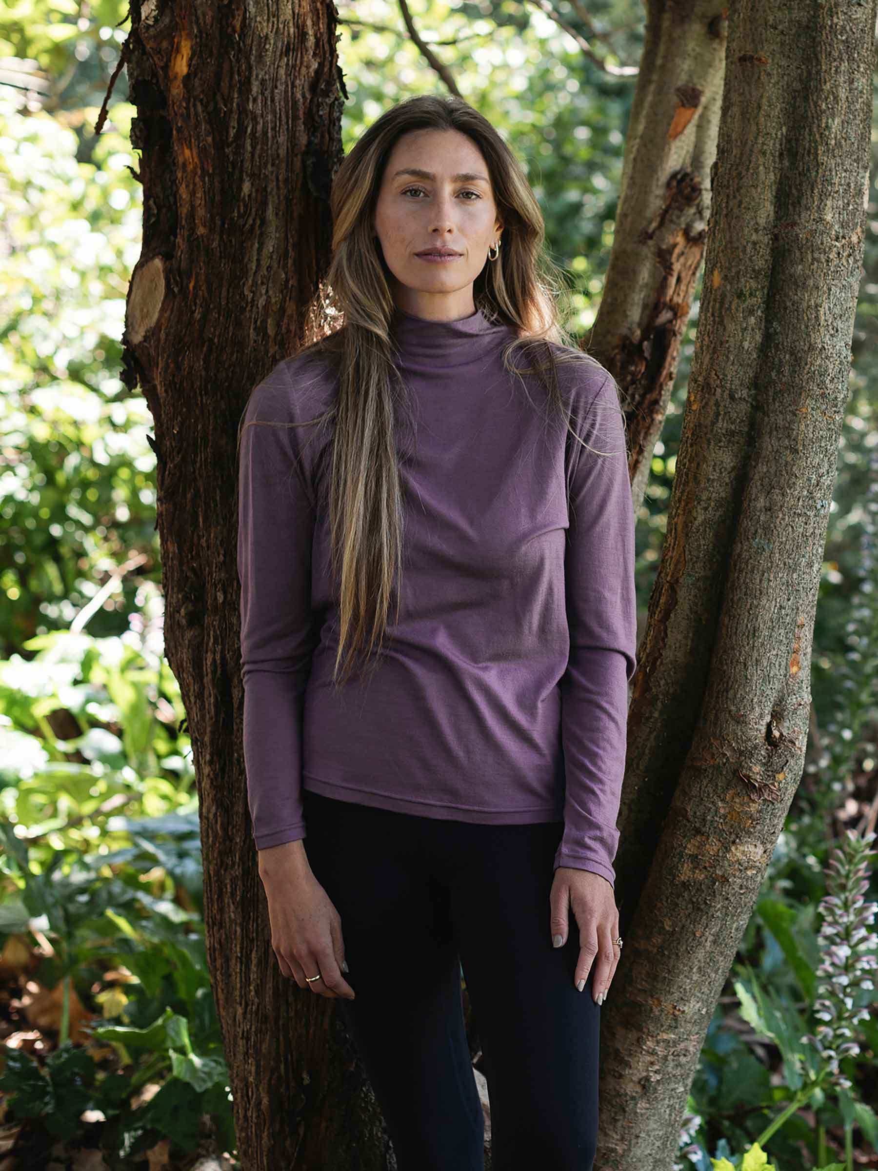 Woman wearing a purple long-sleeve top standing between two trees in a forest.