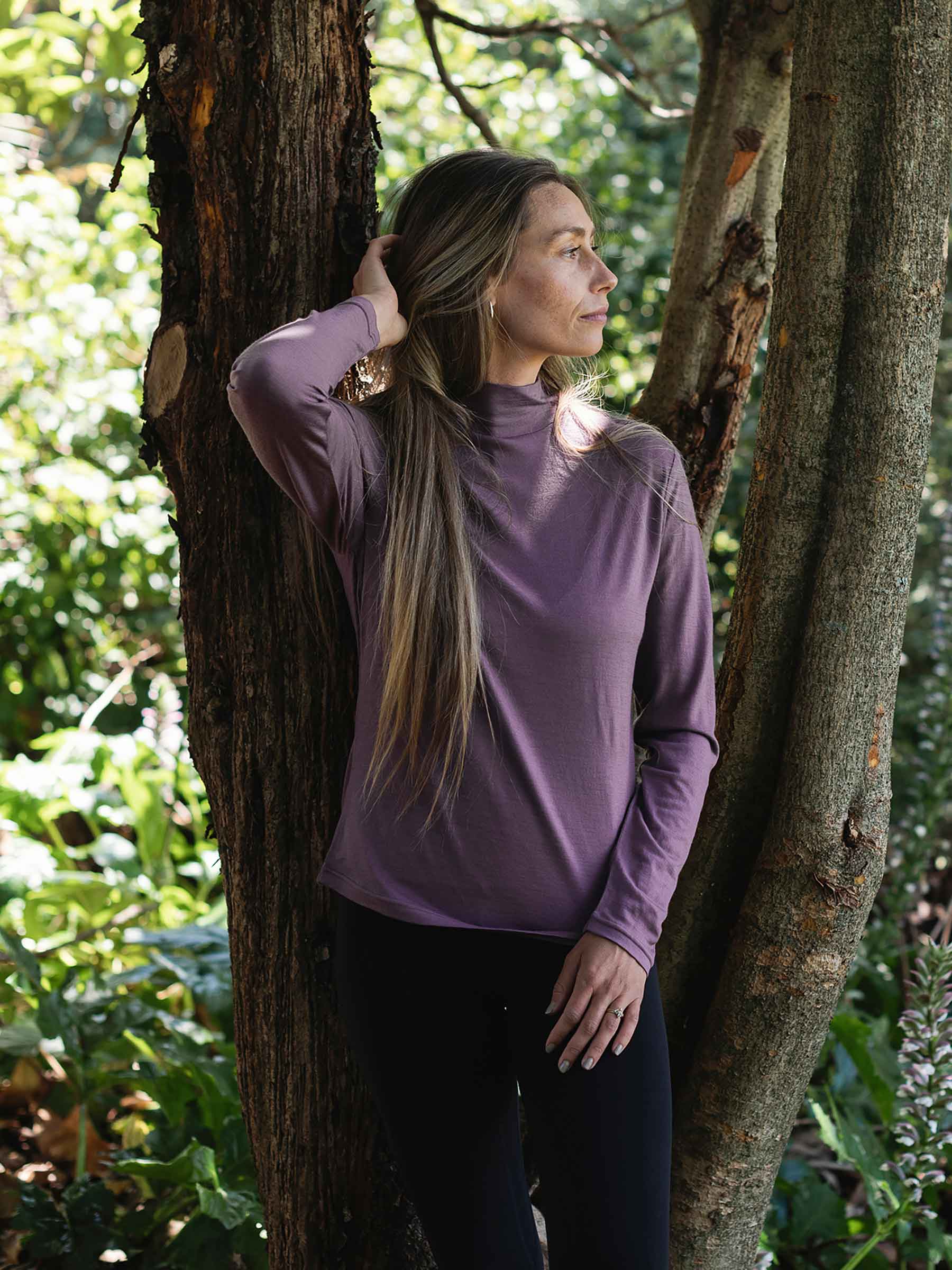 Woman in a purple long-sleeve shirt standing between two trees in a garden.