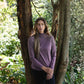 Woman in a purple long-sleeve shirt standing between two trees in a forest.