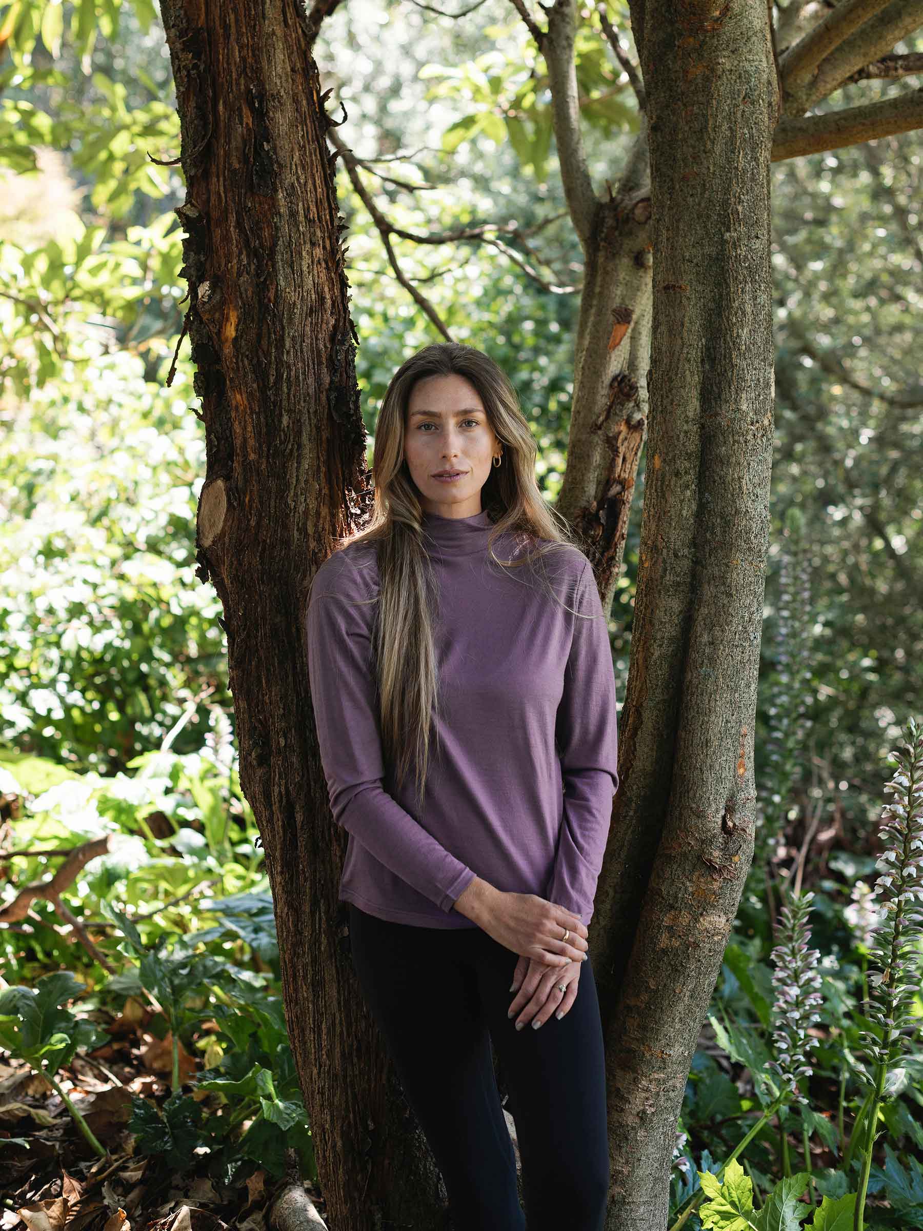 Woman in a purple long-sleeve shirt standing between two trees in a forest.