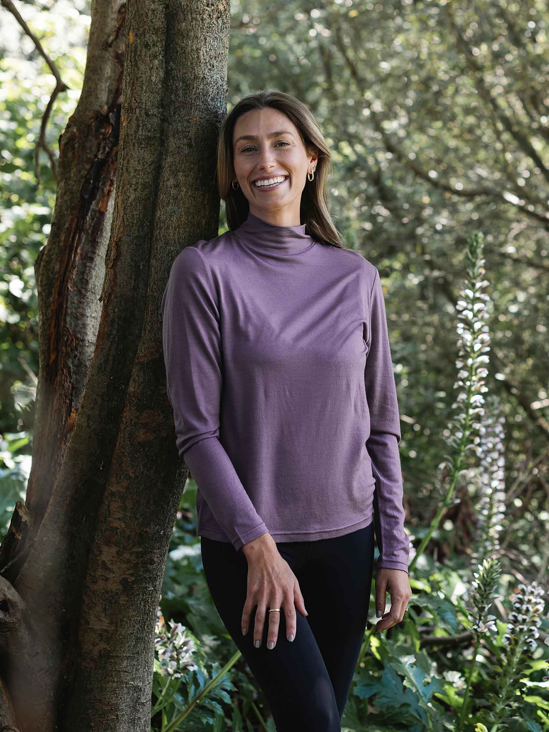 Woman wearing a purple long-sleeve shirt standing next to a tree in a forest
